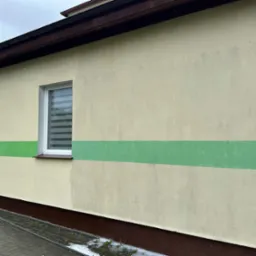 Fragment elewacji budynku z widoczną różnicą w czystości po umyciu fragmentu ściany, z zielonym pasem na górze, szarą ścianą poniżej i widocznym oknem z żaluzjami.