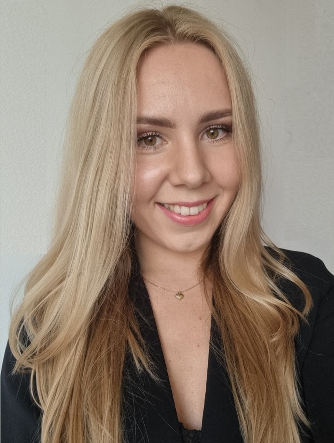 Uśmiechnięta blondynka w czarnej marynarce i delikatnym złotym naszyjniku z zawieszką w kształcie serca, portret na jasnym tle.