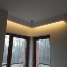 Nowoczesne oświetlenie sufitowe LED w narożniku pokoju z dwoma oknami z brązowymi ramami, widok na drzewa za oknem.