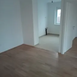 Pusty pok&oacute;j z panelami podłogowymi i świeżo pomalowanymi białymi ścianami, widok na sąsiednie pomieszczenie z oknem i wystającą instalacją elektryczną.