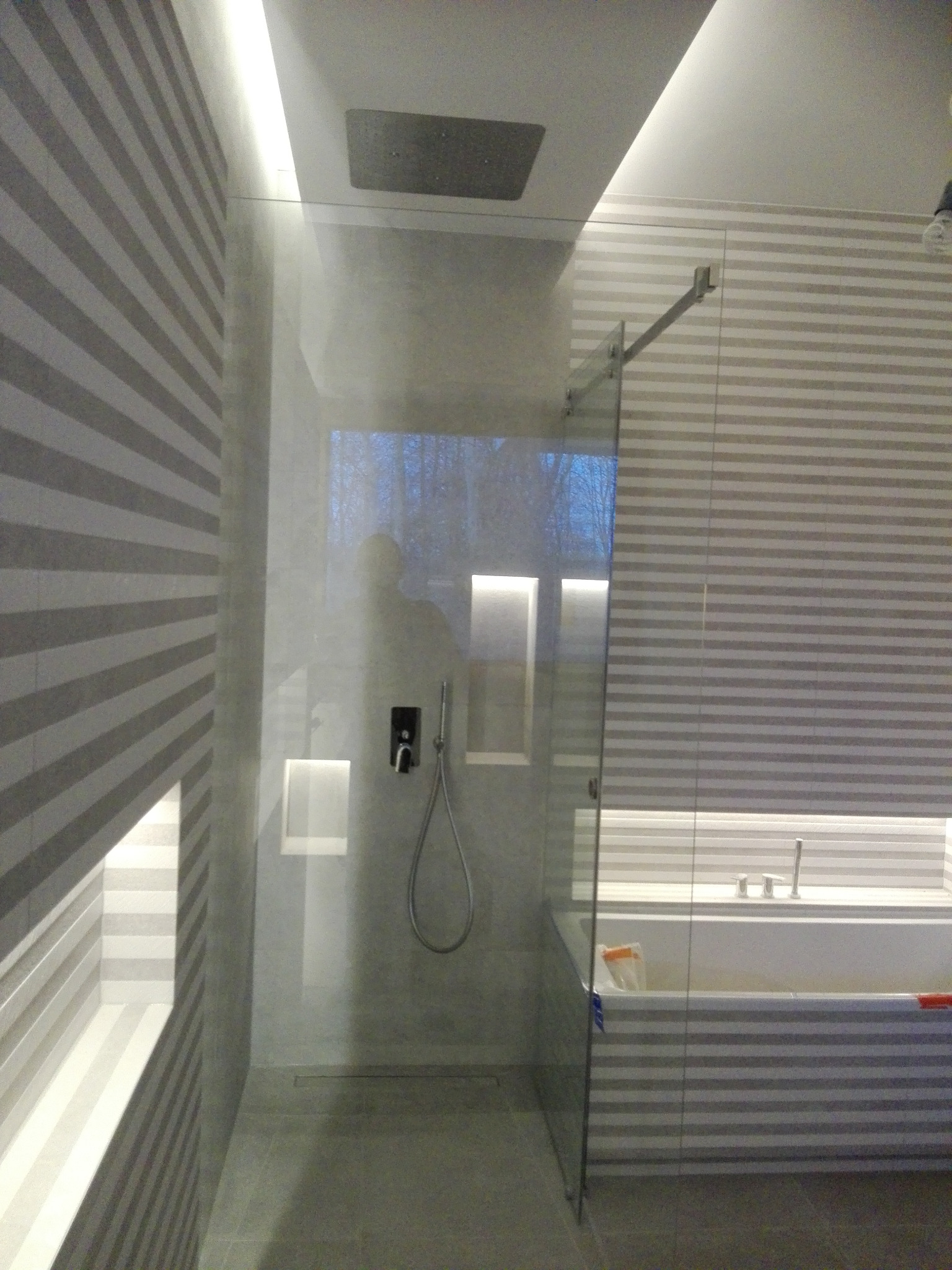 Nowoczesna łazienka z kabiną prysznicową typu walk-in, wykończona płytkami w poziome pasy, z oświetleniem LED w suficie i wnękach, oraz widoczną wanną za szklaną ścianką.