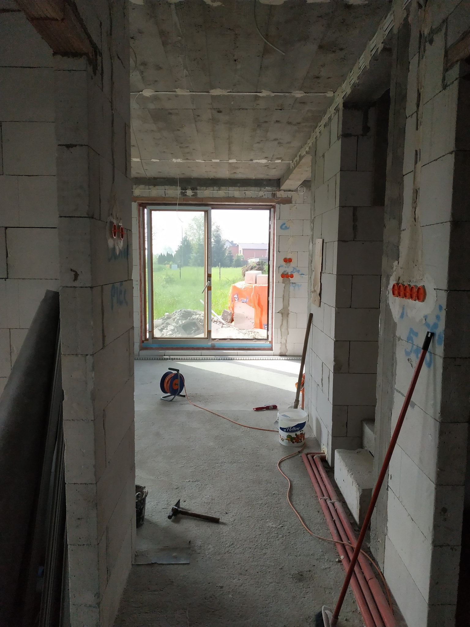 Wnętrze w stanie surowym z widocznymi instalacjami elektrycznymi w ścianach z betonu komórkowego, narzędziami na podłodze i oknem z widokiem na zieleń.