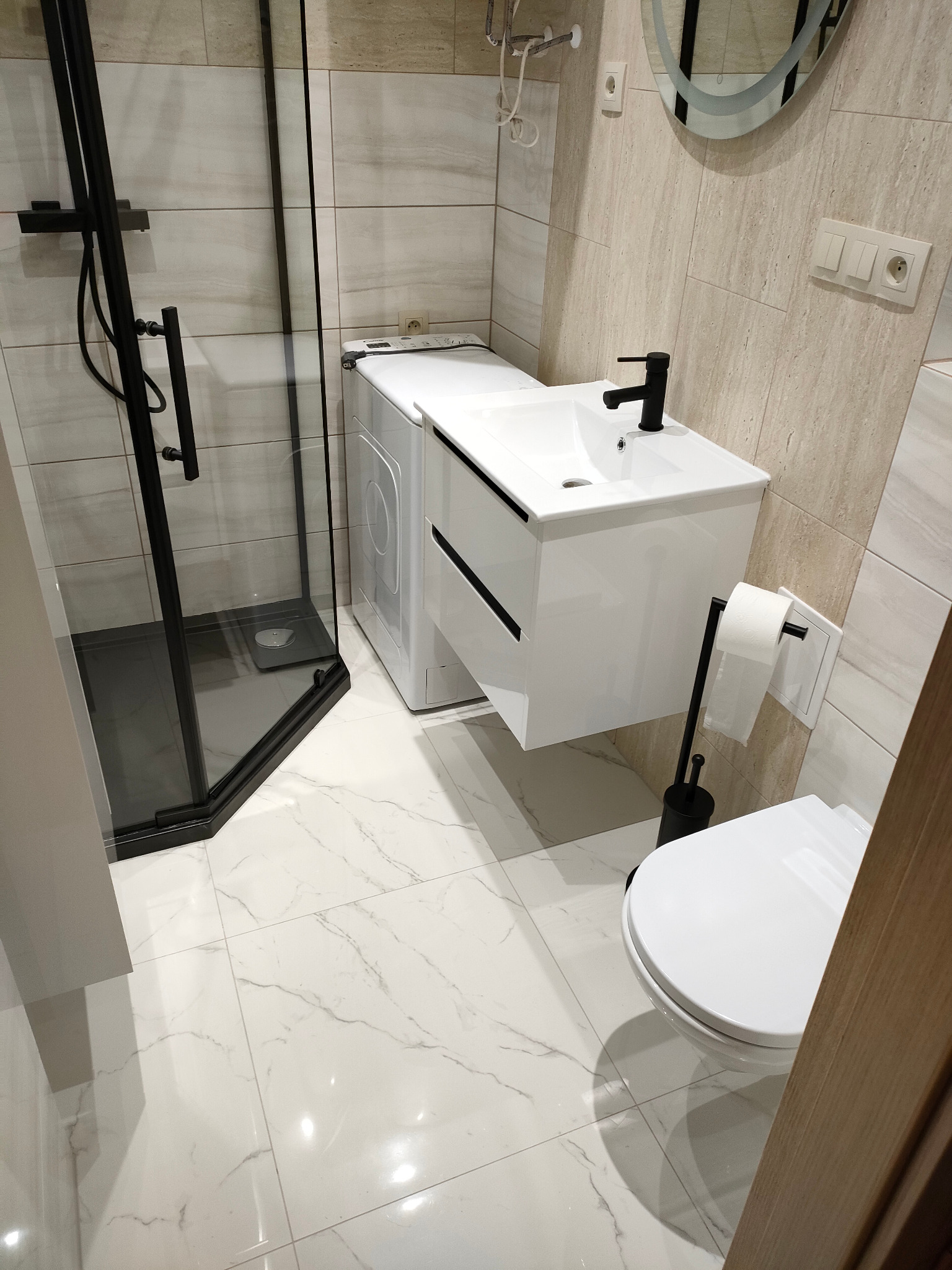Nowoczesna łazienka z kabiną prysznicową typu walk-in z czarnymi elementami, białą umywalką z szafką, toaletą i pralką ukrytą pod blatem, wykończona płytkami w odcieniach beżu i bieli z marmurkowym...