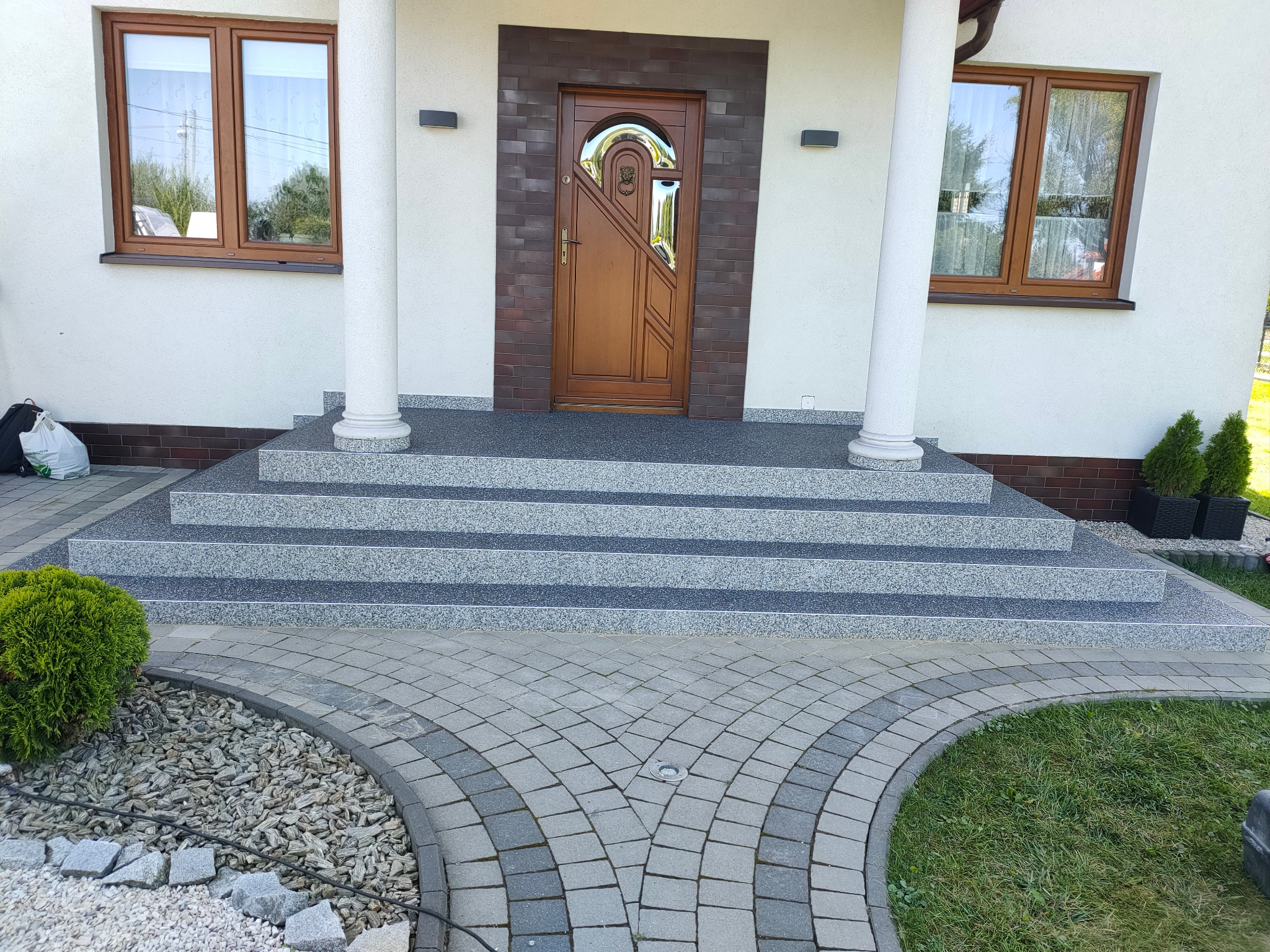 Wejście do domu z czterostopniowymi schodami wyłożonymi szarym kamieniem, brązowe drzwi z szybą i klamką po prawej stronie, ścieżka z szarej kostki brukowej, trawnik, okna z brązowymi ramami.