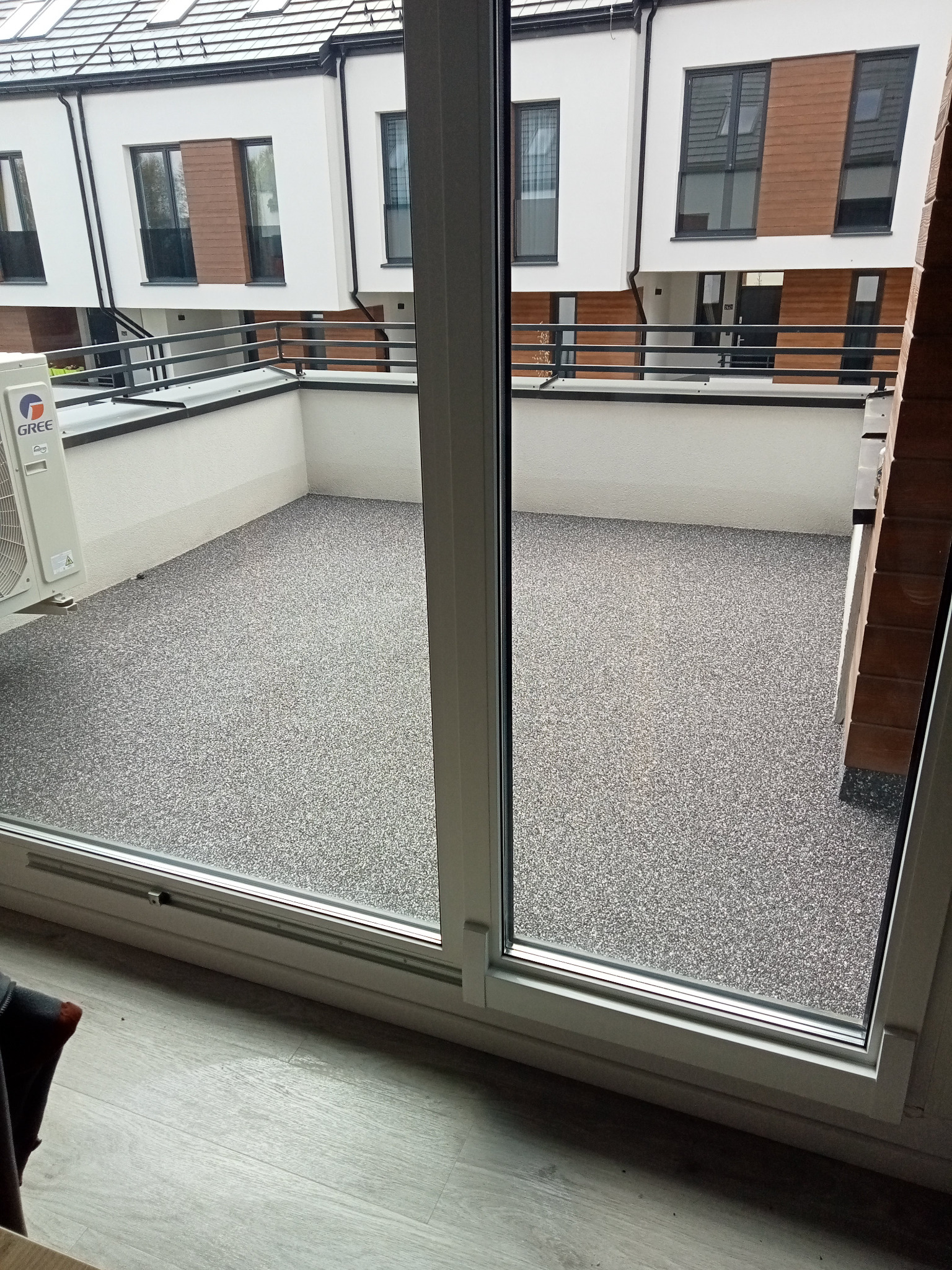 Balkon z szarą, żywiczną posadzką, widok z wnętrza przez okno balkonowe, na zewnątrz widoczne budynki szeregowe z elementami drewnianymi.
