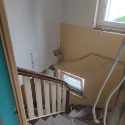 RAFBUD RAFAŁ STYPIŃSKI - Klatka schodowa w trakcie remontu: widoczne schody, barierka z drewnianym poręczą i białymi tralkami, ściany częściowo pomalowane na żółto, okno z widoczną pianką montażową, wystające kable.