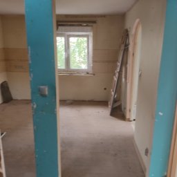 RAFBUD RAFAŁ STYPIŃSKI - Wnętrze pokoju w trakcie remontu, widoczna drabina oparta o ścianę, surowe ściany, okno z widokiem na zieleń, turkusowa framuga drzwi z widocznymi śladami farby.