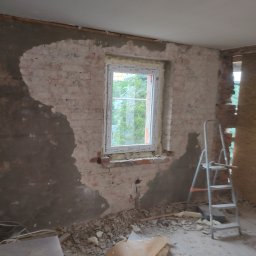 RAFBUD RAFAŁ STYPIŃSKI - Wnętrze w trakcie remontu: fragment ściany z oknem, częściowo otynkowana cegła, widoczne ślady po kuciu, gruz na podłodze i drabina aluminiowa.