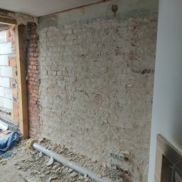 RAFBUD RAFAŁ STYPIŃSKI - Ceglana ściana w trakcie remontu, częściowo otynkowana, z widocznymi instalacjami elektrycznymi i rurą kanalizacyjną na podłodze zasypanej gruzem.