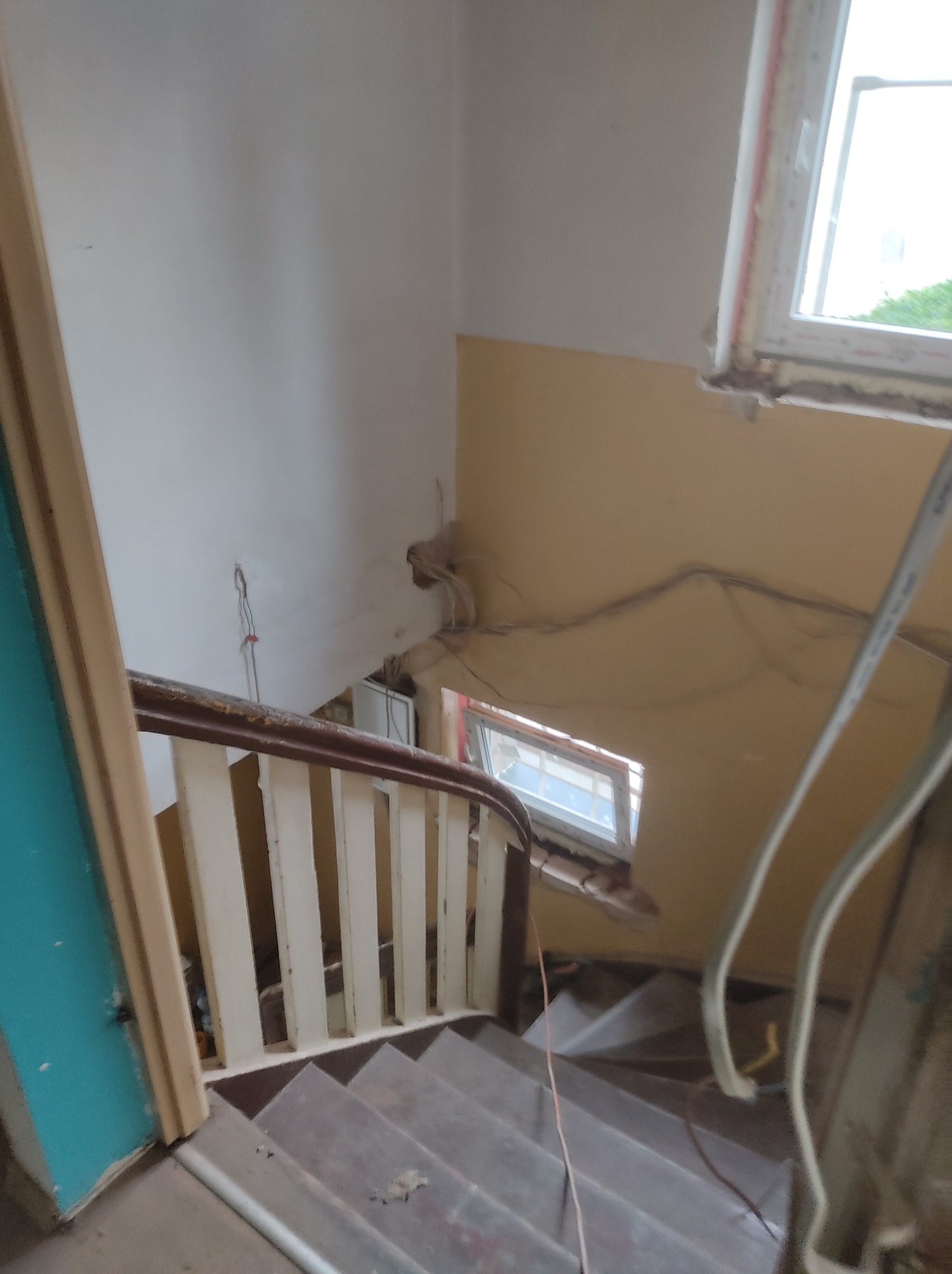 Klatka schodowa w trakcie remontu: widoczne schody, barierka z drewnianym poręczą i białymi tralkami, ściany częściowo pomalowane na żółto, okno z widoczną pianką montażową, wystające kable.