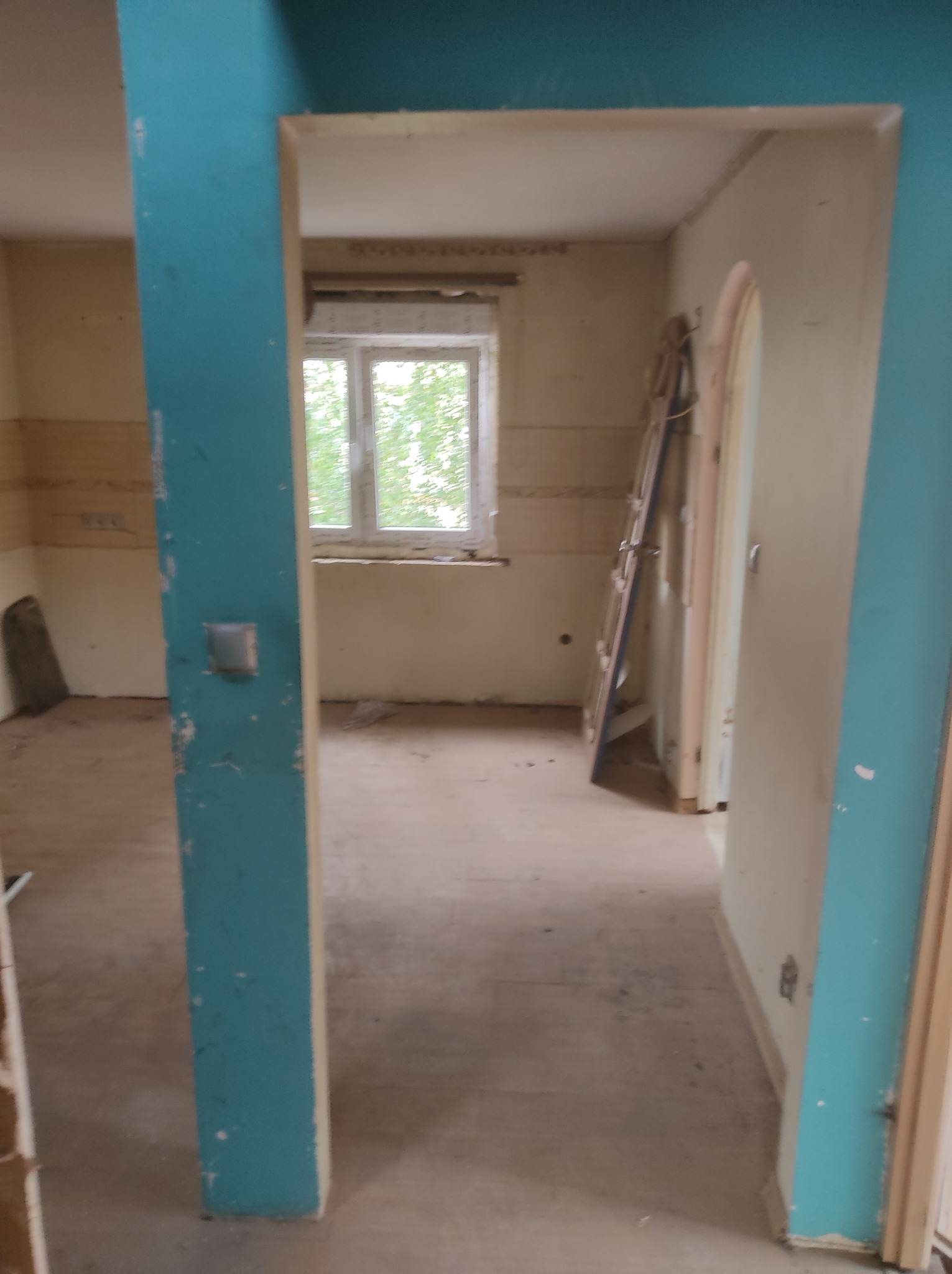 Wnętrze pokoju w trakcie remontu, widoczna drabina oparta o ścianę, surowe ściany, okno z widokiem na zieleń, turkusowa framuga drzwi z widocznymi śladami farby.