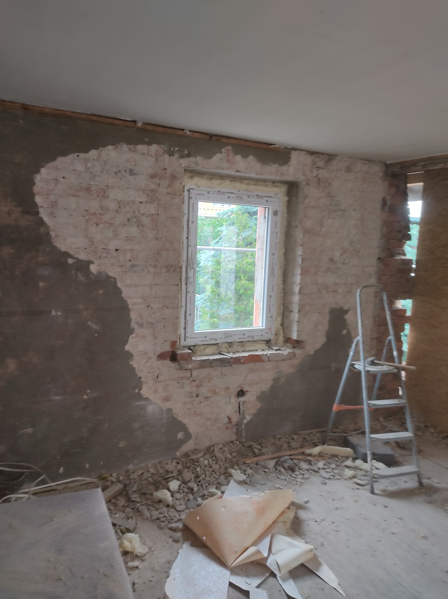 Wnętrze w trakcie remontu: fragment ściany z oknem, częściowo otynkowana cegła, widoczne ślady po kuciu, gruz na podłodze i drabina aluminiowa.