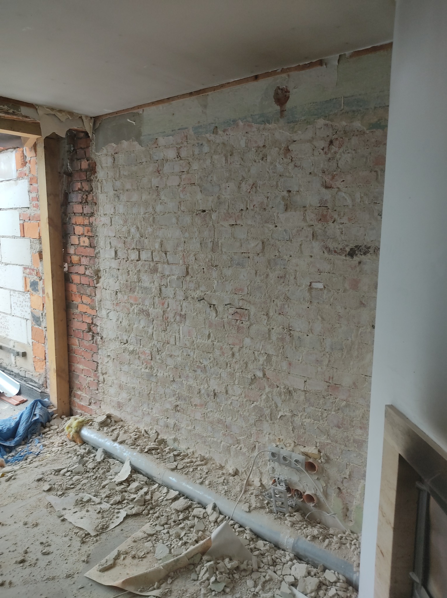 Ceglana ściana w trakcie remontu, częściowo otynkowana, z widocznymi instalacjami elektrycznymi i rurą kanalizacyjną na podłodze zasypanej gruzem.