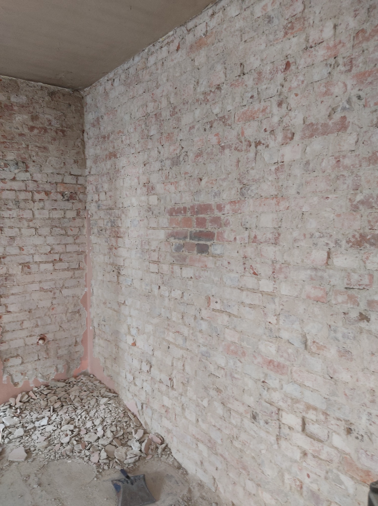 Odsłonięta ceglana ściana w trakcie remontu, widoczne resztki gruzu i zaprawy na podłodze, fragment ściany z ciemniejszej cegły.