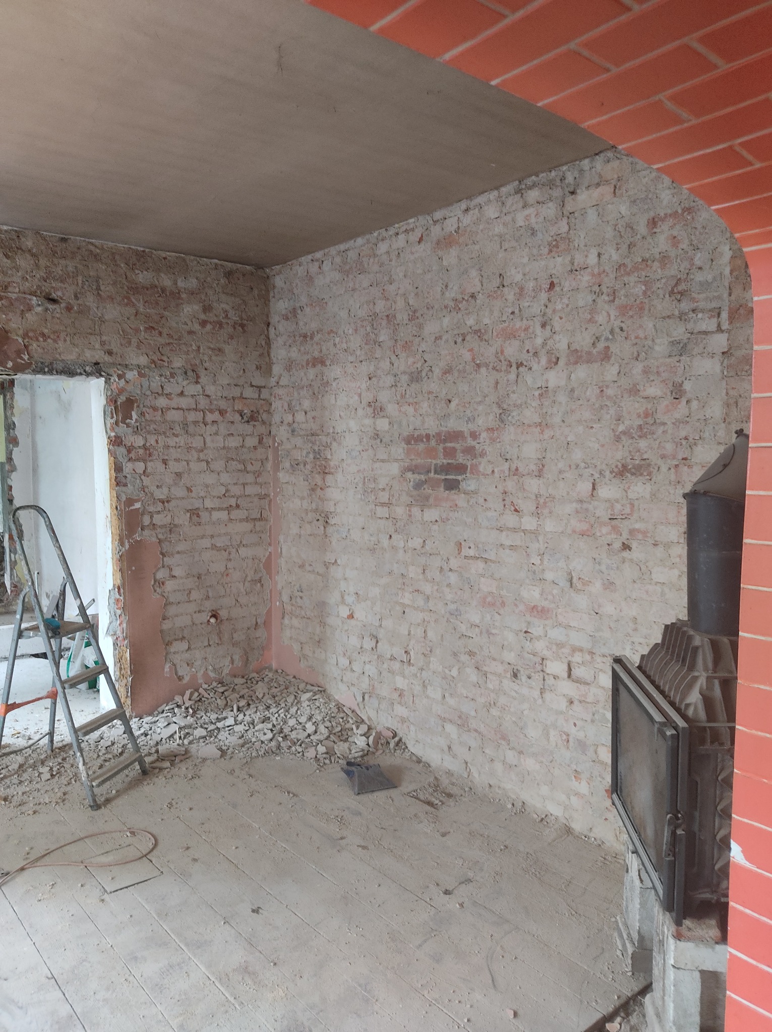 Pokój w trakcie remontu: odsłonięta cegła na ścianach, częściowo otynkowany sufit, drabina aluminiowa w rogu, gruz na podłodze, widoczny piec kaflowy z otwartymi drzwiczkami.