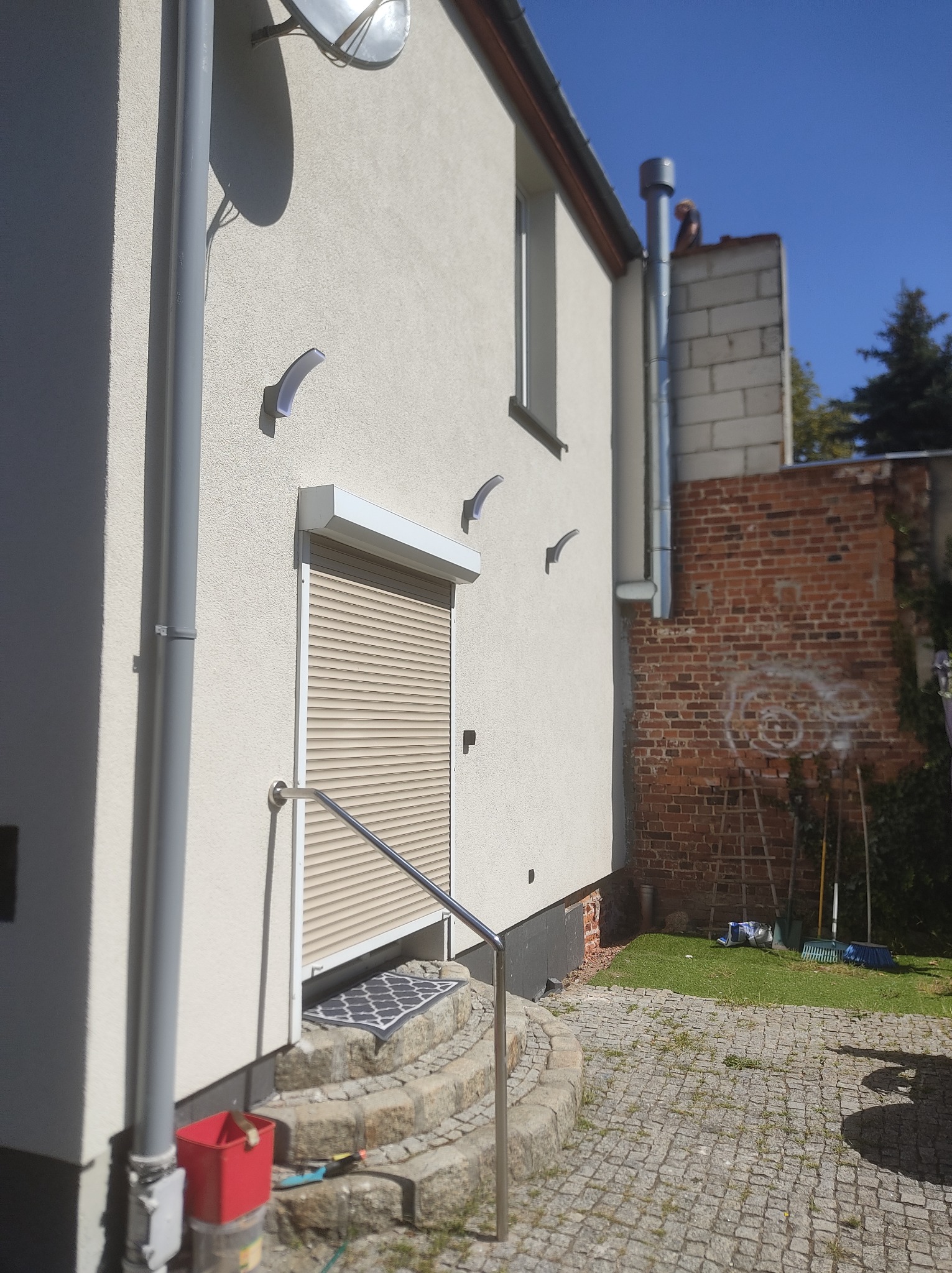 Jasna elewacja domu z beżowymi roletami zewnętrznymi, widoczne schody wejściowe z metalową barierką, oświetlenie elewacyjne i elementy instalacji odgromowej.