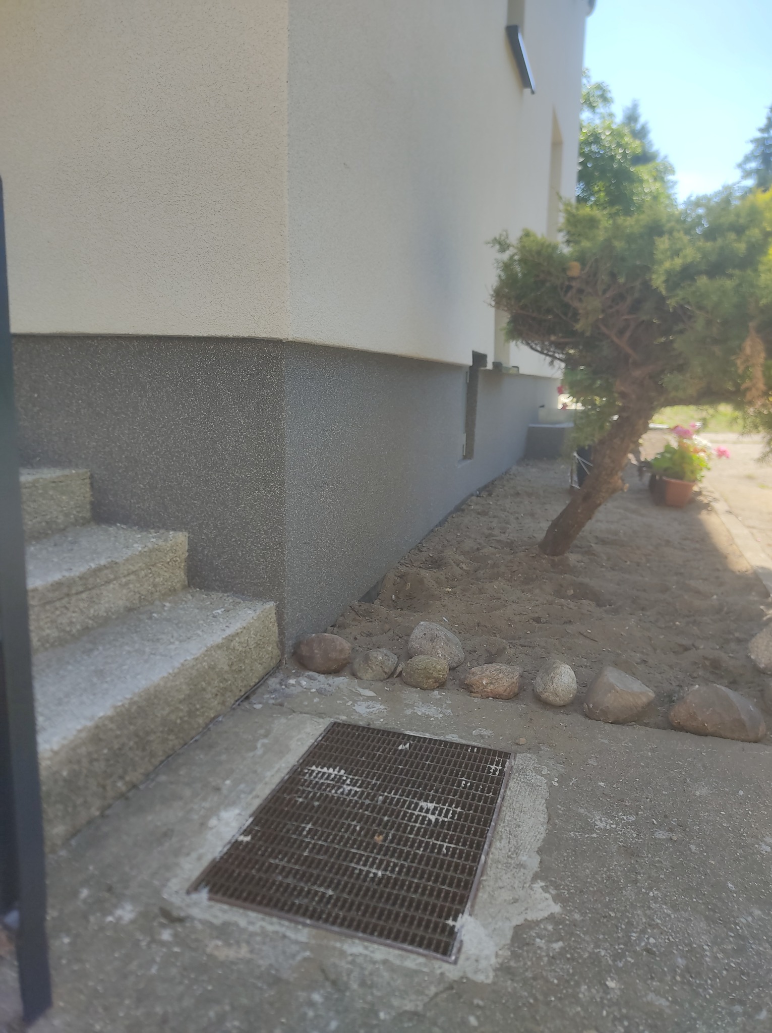 Wykończona elewacja domu z widocznym cokołem w szarym kolorze, schodami wejściowymi i rabatą z kamieniami oraz kratką ściekową.