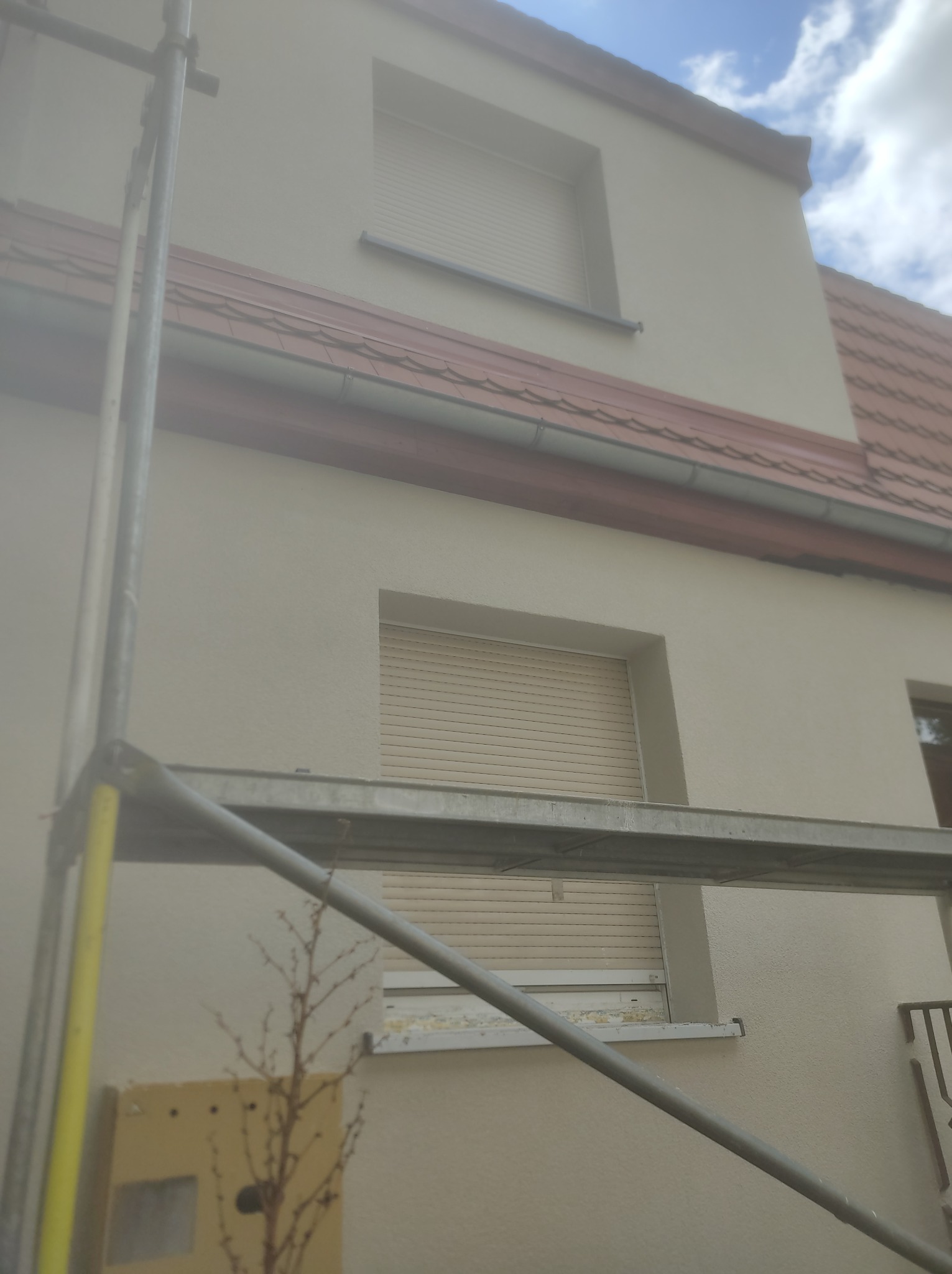 Elewacja budynku jednorodzinnego w trakcie ocieplania, widoczne rusztowanie z żółtymi elementami, okna z opuszczonymi roletami, fragment dachu z czerwonej dachówki.