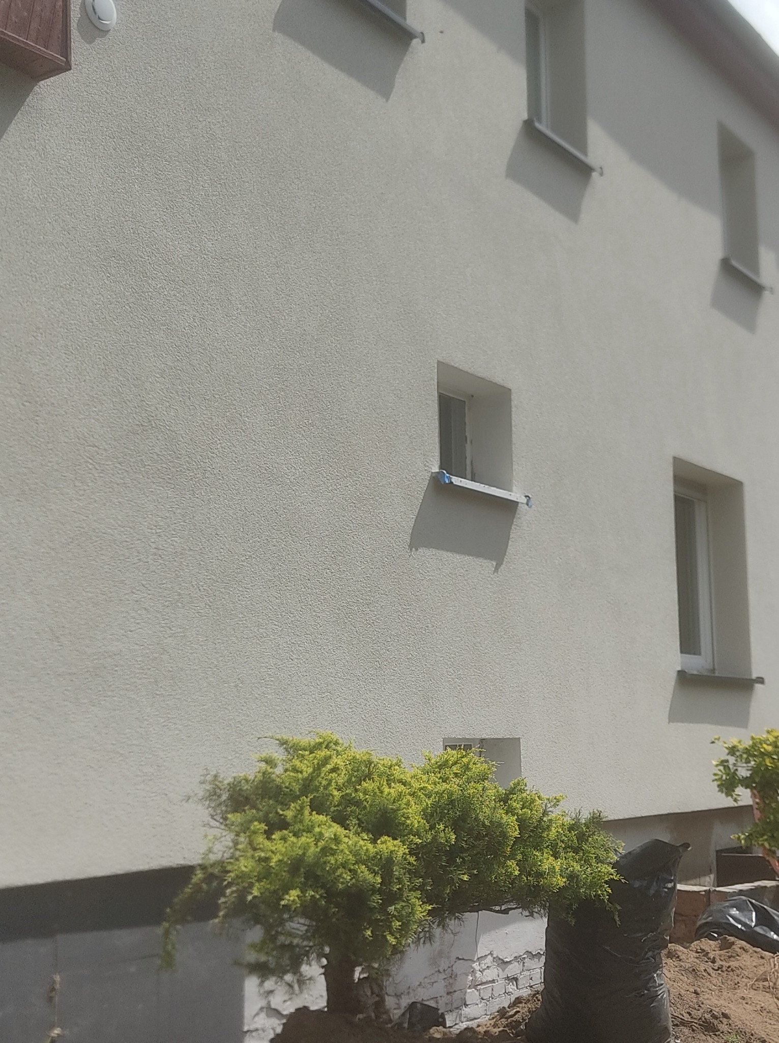 Jasna elewacja budynku z widoczną strukturą, okna z taśmą zabezpieczającą, krzewy iglaste przy fundamencie, czarne worki z ziemią.