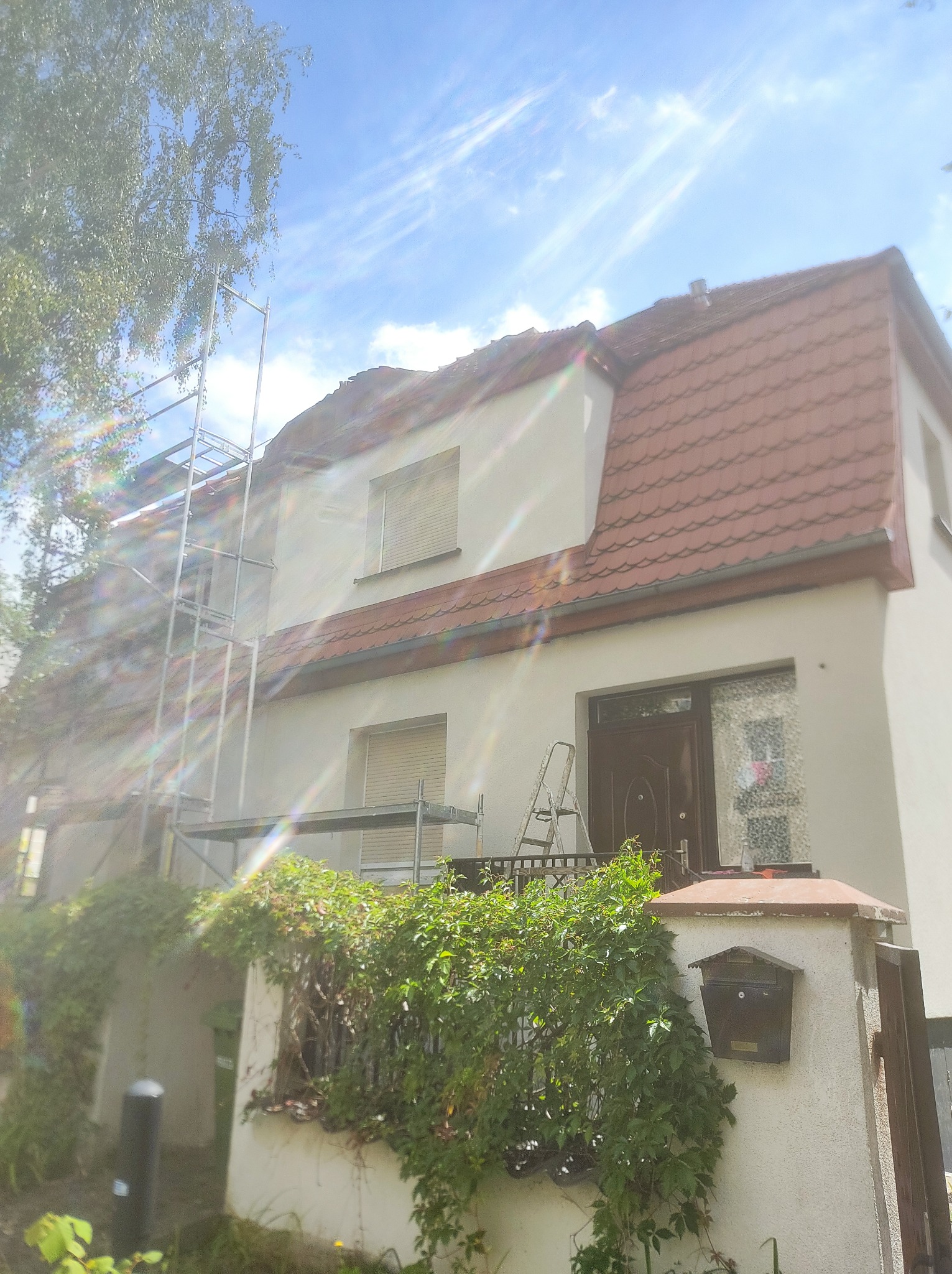 Dwupiętrowy dom z czerwonym dachem w trakcie prac elewacyjnych, widoczne rusztowania i drabina, fragment ogrodzenia porośniętego zielenią, niebieskie niebo z chmurami w tle.