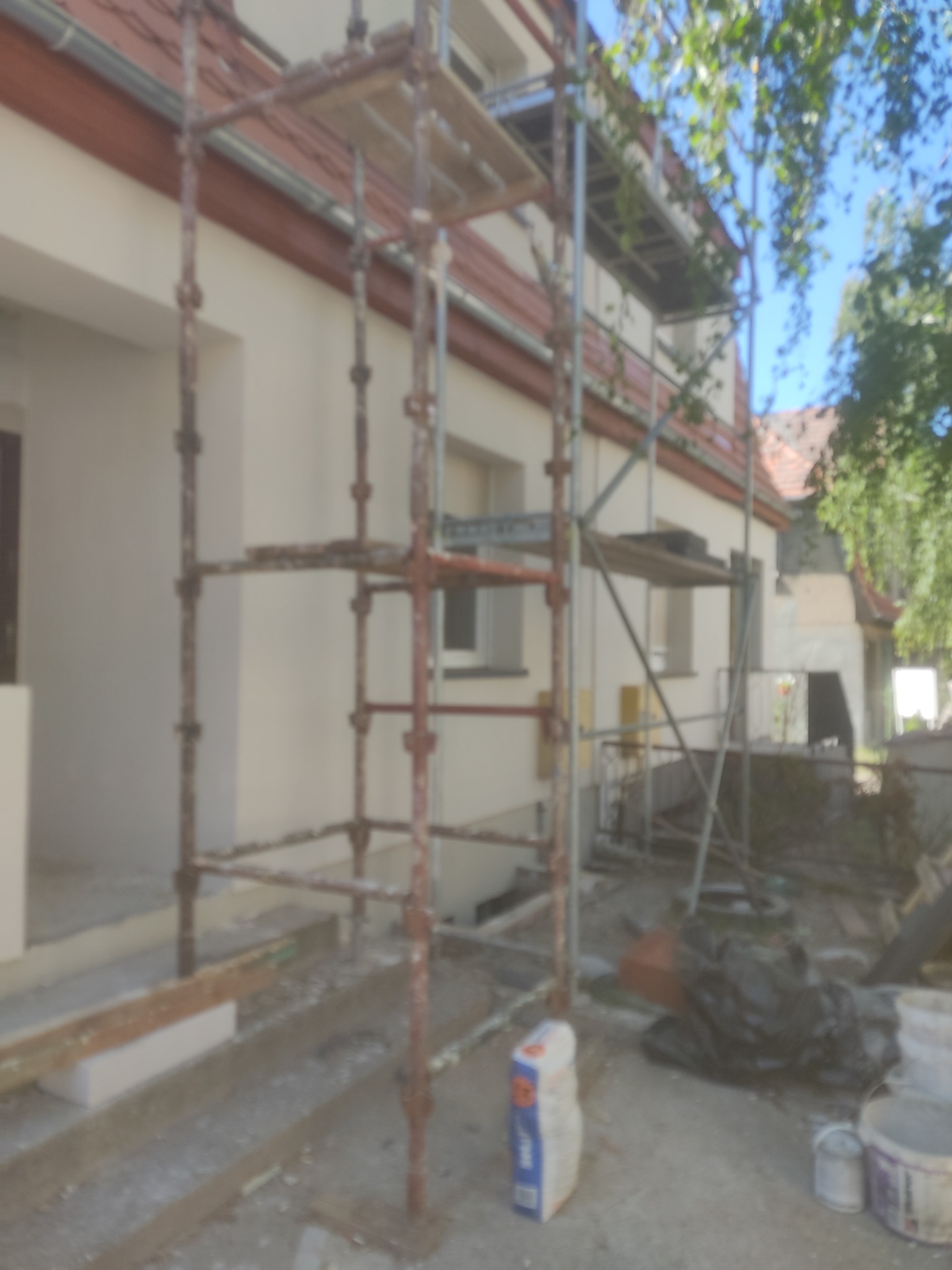 Dom jednorodzinny w trakcie ocieplania elewacji, widoczne rusztowanie, worki z zaprawą i narzędzia budowlane na zewnątrz.