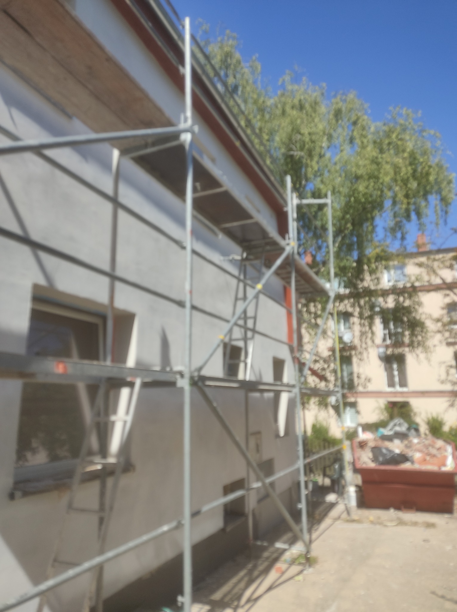 Dom w trakcie prac elewacyjnych, widoczne rusztowania, ściana pokryta szarym tynkiem, okna zabezpieczone, w tle kontener na gruz i budynki mieszkalne.