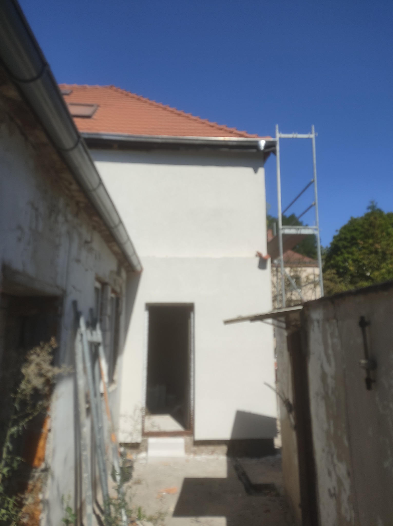 Świeżo otynkowana ściana budynku z widocznym rusztowaniem i fragmentami starej elewacji, zarośnięte chwasty u podstawy ściany.