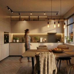 Julia Dujka Interior Design - Nowoczesna kuchnia z jadalnią: minimalistyczne szafki, drewniany stół z owocami, dekoracyjne oświetlenie i regał na wino z podświetleniem LED. Kobieta w tle.