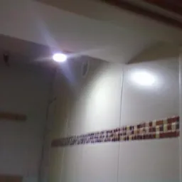 Wykończona łazienka z białymi płytkami i dekoracyjnym pasem mozaiki. Widoczne oświetlenie punktowe na suficie i fragment ramy drzwi.