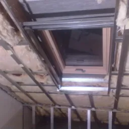 Montaż okna dachowego w skośnym dachu z izolacją z wełny mineralnej, widoczne metalowe profile konstrukcyjne. Etap prac wykończeniowych poddasza.