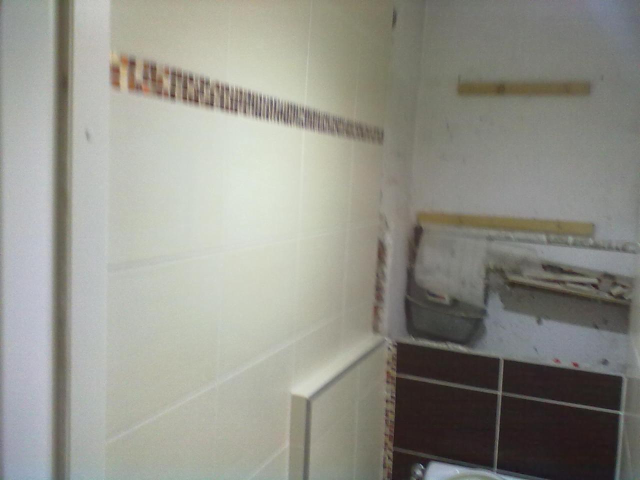 Wnętrze łazienki w trakcie remontu. Widoczne białe płytki na ścianach z dekoracyjnym paskiem mozaiki i przygotowane miejsce pod zabudowę umywalki. Ściany częściowo bez wykończenia.