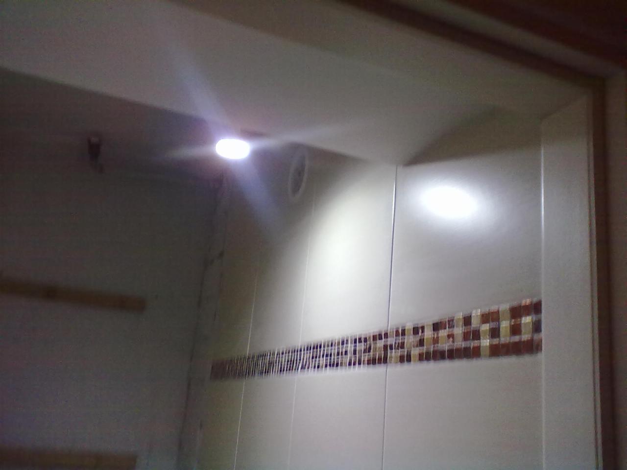 Wykończona łazienka z białymi płytkami i dekoracyjnym pasem mozaiki. Widoczne oświetlenie punktowe na suficie i fragment ramy drzwi.