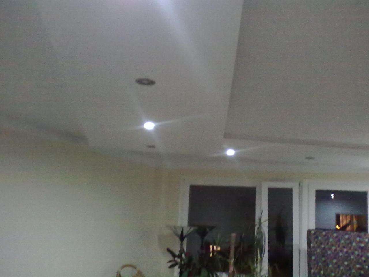 Jasne wnętrze z podwieszanym sufitem i oświetleniem punktowym. Widoczne drzwi balkonowe i fragment ściany. Neutralna kolorystyka.