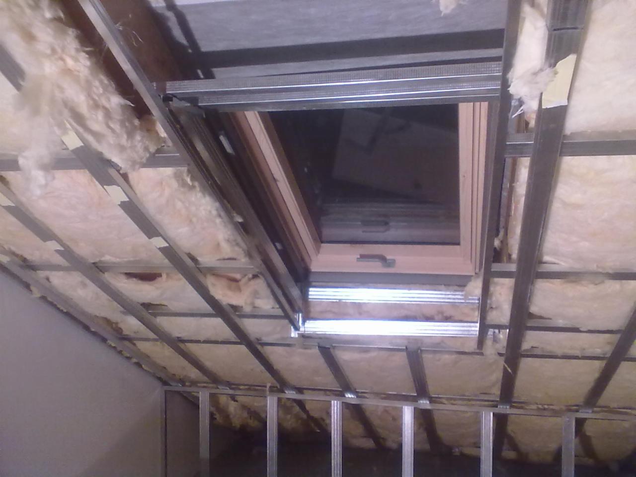 Montaż okna dachowego w skośnym dachu z izolacją z wełny mineralnej, widoczne metalowe profile konstrukcyjne. Etap prac wykończeniowych poddasza.