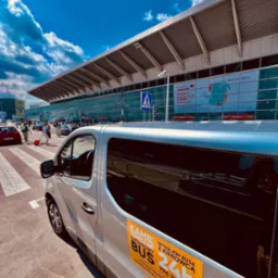 Srebrny bus z reklamą wynajmu z kierowcą, zaparkowany przed lotniskiem w słoneczny dzień, widoczni przechodnie na pasach.