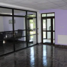 Wnętrze biurowe z przeszkloną ścianą, widoczne biurko z krzesłem, podłoga wyłożona ciemnymi płytkami, ściana pomalowana na fioletowo, biały grzejnik