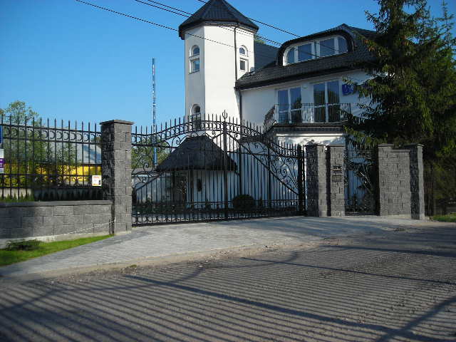 Dwupiętrowy budynek biurowy z wieżyczką, widoczny zza ozdobnego, kutego ogrodzenia z kamiennymi słupkami. Na elewacji widoczne logo firmy.