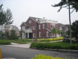 Dwupiętrowy dom z czerwonej cegły z białymi oknami i balkonem, otoczony niskimi krzewami i drzewami, z innym domem widocznym w tle, sfotografowany w pochmurny dzień.