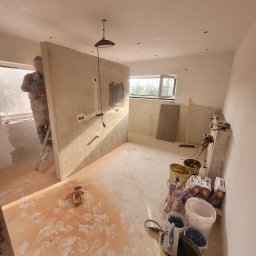 USŁUGI OGÓLNOBUDOWLANE FILIP HOJA - Wnętrze w trakcie prac wykończeniowych: montaż płyty kartonowo-gipsowej z przygotowanymi otworami na instalacje elektryczne, widoczne ślady obuwia na podłodze, wiadra z zaprawą i narzędzia w rogu...