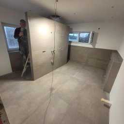 USŁUGI OGÓLNOBUDOWLANE FILIP HOJA - Montaż płytek wielkoformatowych w łazience; widoczny glazurnik na drabinie z szlifierką kątową, docięte płytki na ścianie z wyprowadzonymi instalacjami, szare płytki podłogowe.