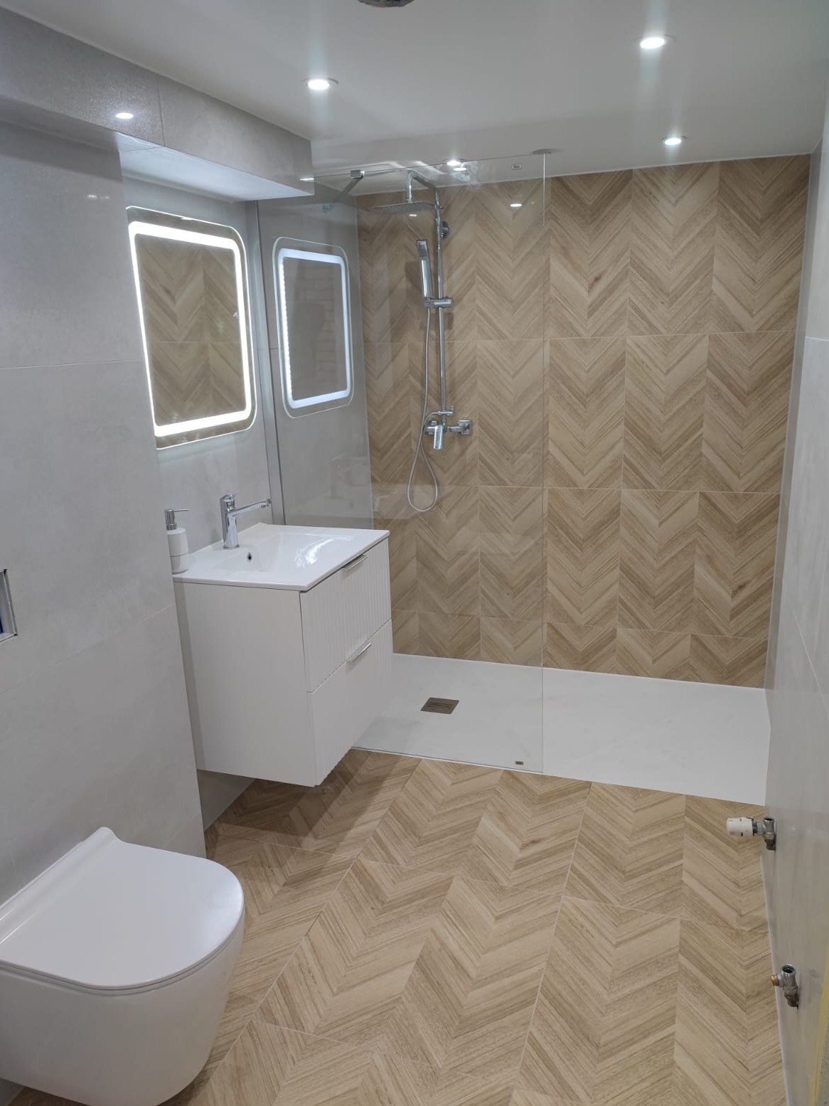 Nowoczesna łazienka z beżowymi płytkami ułożonymi w jodełkę na podłodze i ścianie prysznica, biała szafka z umywalką i podświetlane lustra.