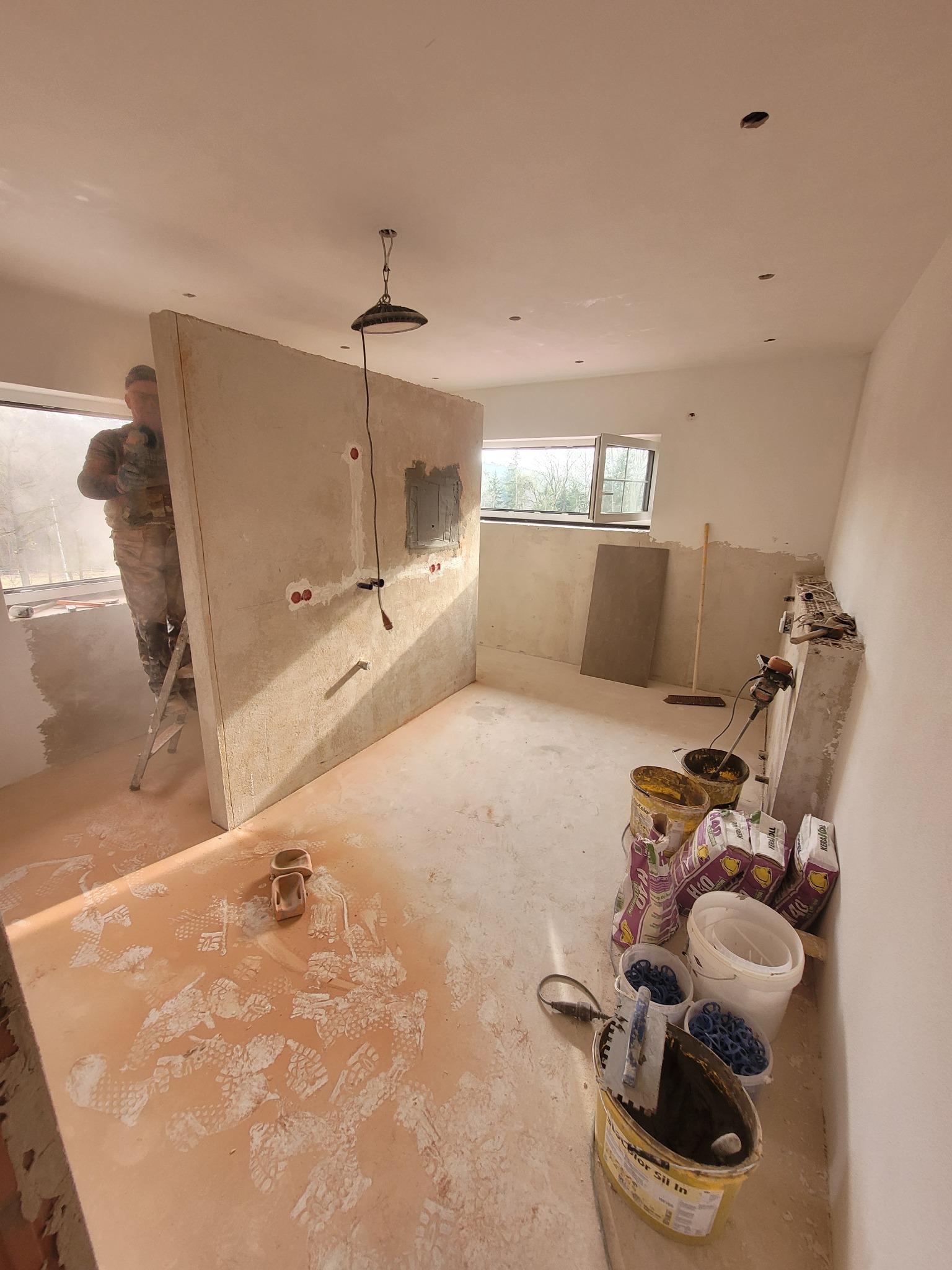 Wnętrze w trakcie prac wykończeniowych: montaż płyty kartonowo-gipsowej z przygotowanymi otworami na instalacje elektryczne, widoczne ślady obuwia na podłodze, wiadra z zaprawą i narzędzia w rogu...