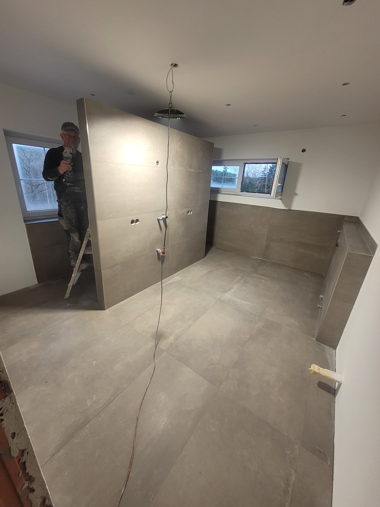 Montaż płytek wielkoformatowych w łazience; widoczny glazurnik na drabinie z szlifierką kątową, docięte płytki na ścianie z wyprowadzonymi instalacjami, szare płytki podłogowe.