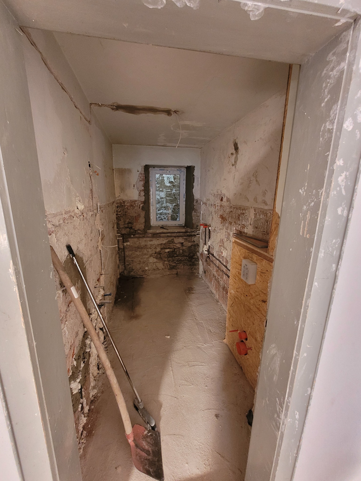 Wnętrze pomieszczenia w trakcie prac przygotowawczych do remontu: odsłonięta cegła, instalacje hydrauliczne, łopaty opierające się o ścianę, okno z widokiem na ceglany mur.