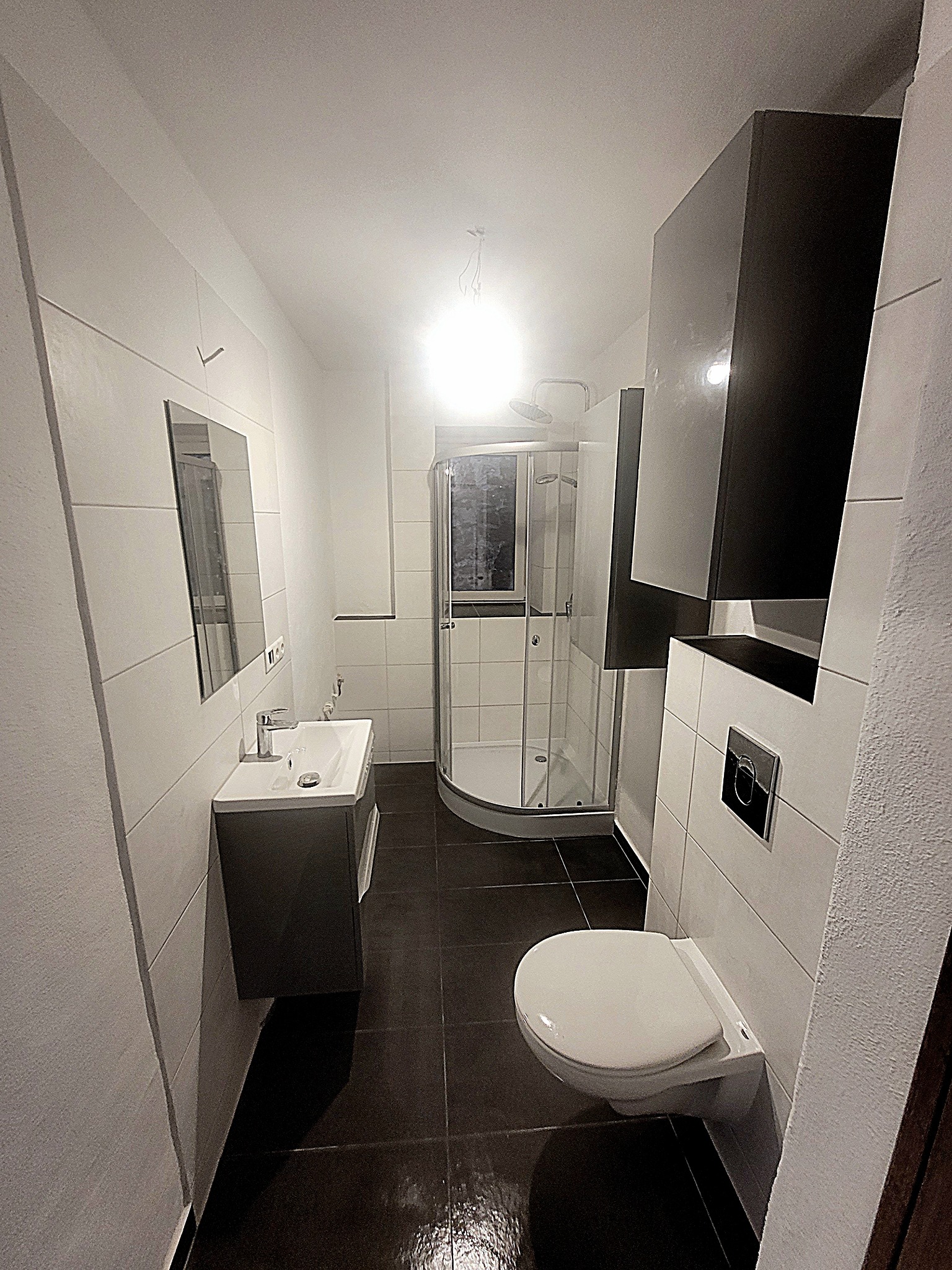 Wykończona łazienka z kabiną prysznicową, wiszącą toaletą, umywalką z szafką i ciemną podłogą, oświetlona przez górne światło.