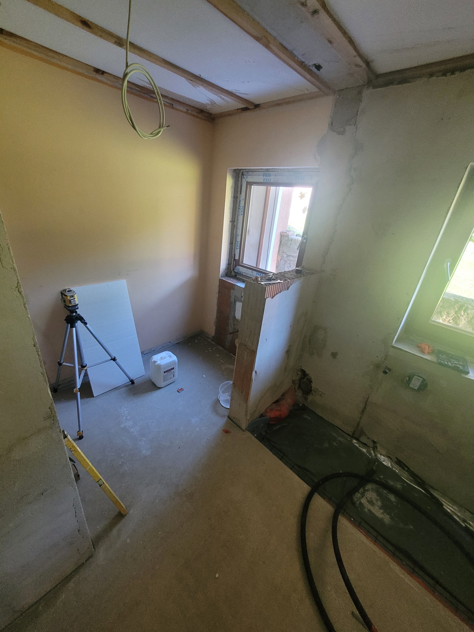 Pokój w trakcie remontu: surowe ściany, odsłonięta konstrukcja sufitu, laser krzyżowy na statywie, płyta karton-gips i poziomica przy ścianie, okno z częściowo obrobionym murem.