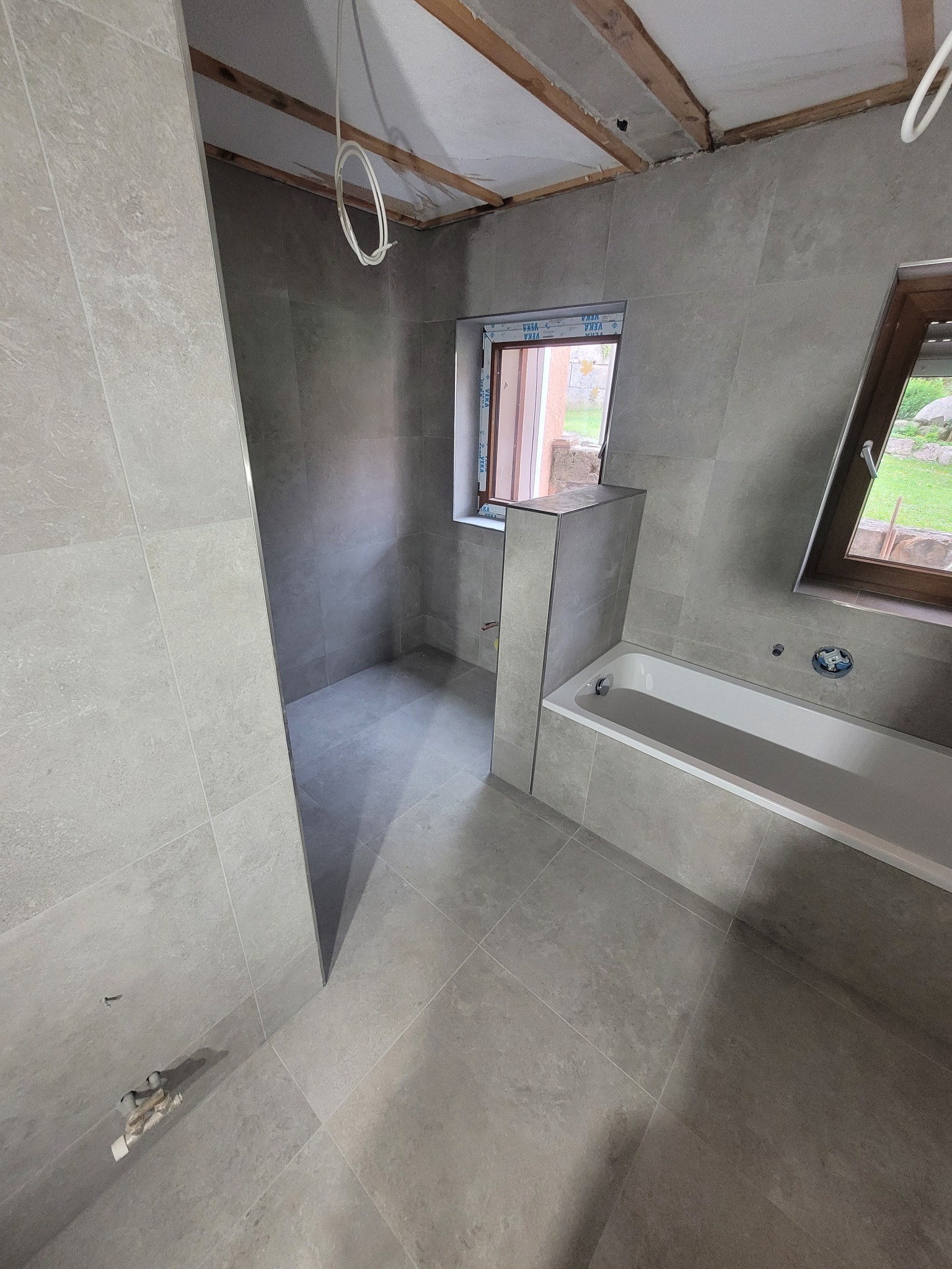 Łazienka w trakcie remontu, wyłożona szarymi płytkami, widoczna wanna, dwa okna z widokiem na zieleń, niedokończony sufit z wystającymi kablami.