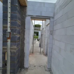 Wnętrze budowy: ściany z białych bloczków, podparte metalowymi stemplami. Widok przez otwory drzwiowe na dalszą część konstrukcji i zieleń. Etap murowania ścian.