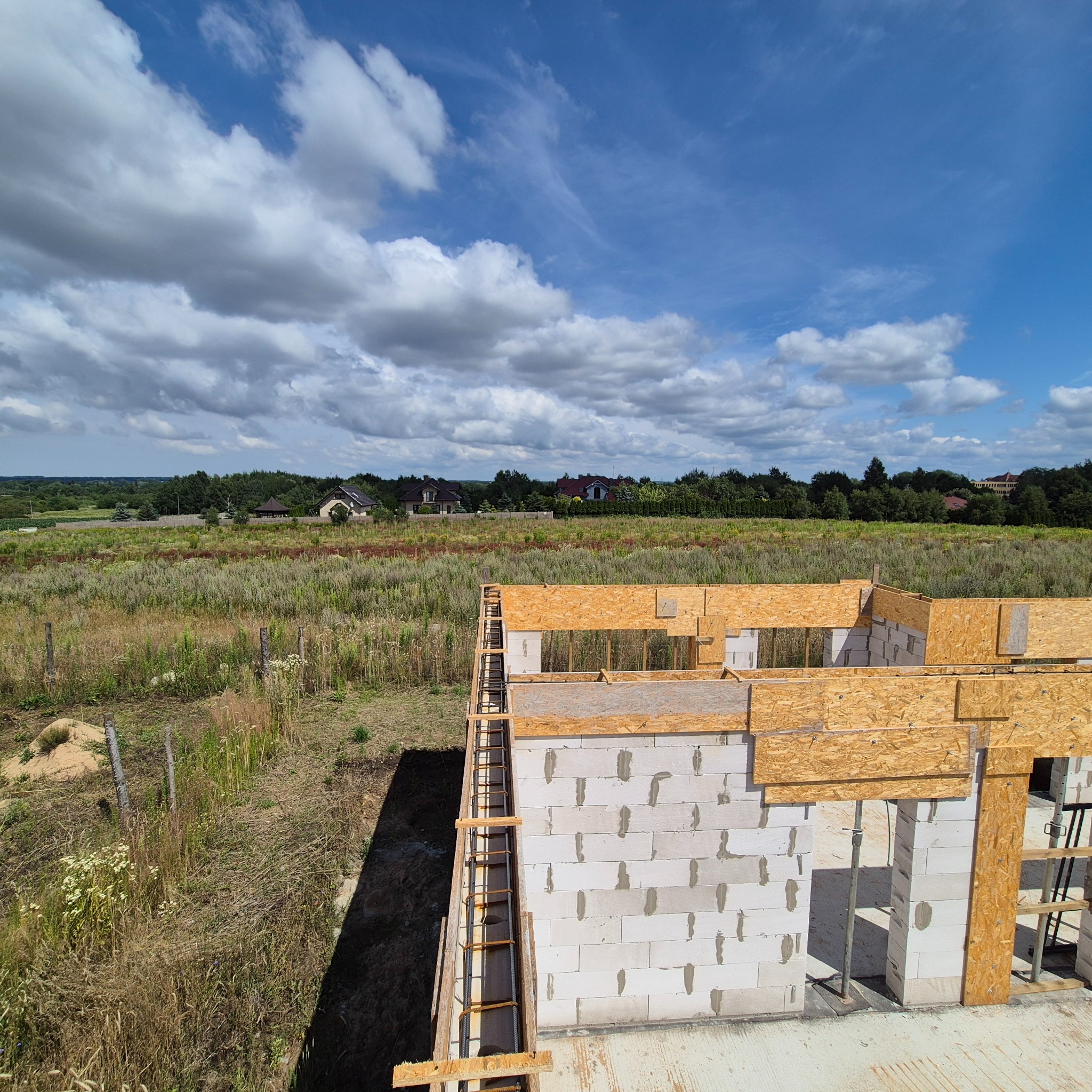 Wysoki kąt: budowa domu z białych bloczków, widoczne szalunki i zbrojenia. W tle łąka i domy pod błękitnym niebem z chmurami.