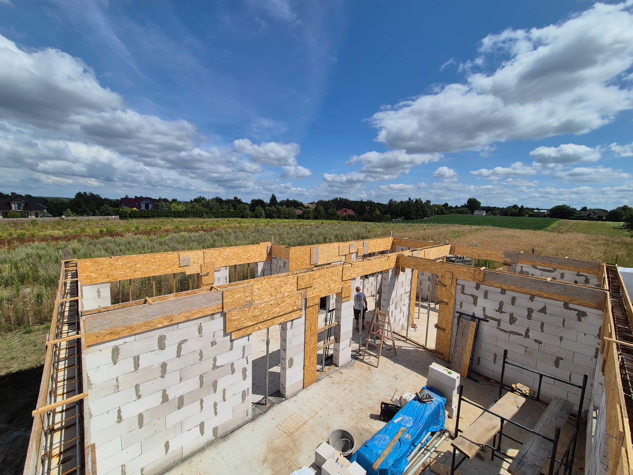 Budowa domu w toku: białe ściany z betonu komórkowego, drewniane szalunki, widok z góry na tle błękitnego nieba z chmurami i zielonych pól. Robocza atmosfera.